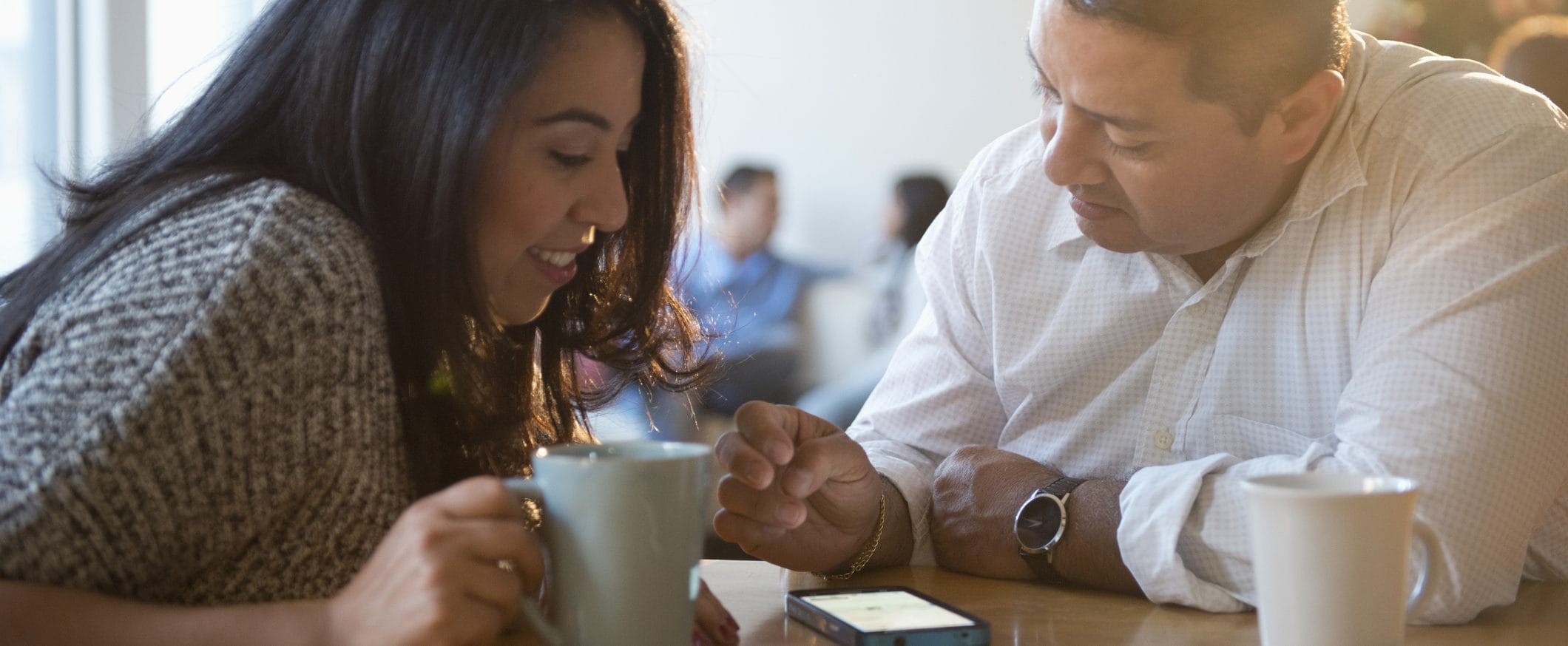 A man and woman lean over a table looking at the screen of a smart phone. The woman holds a coffee mug, and the man has a mug beside him.