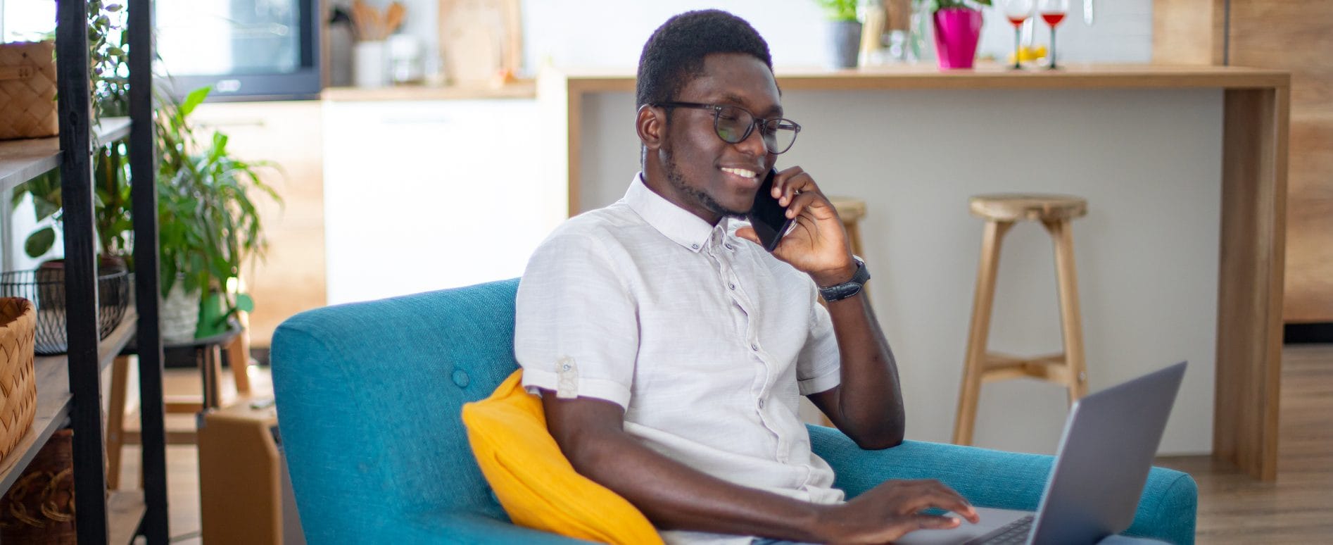 A man sits in an armchair with his laptop computer and talks on a mobile phone.