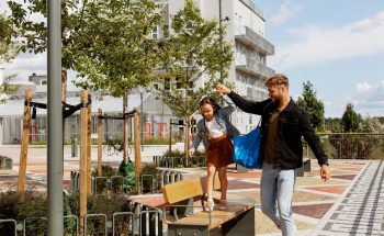 A father holds his daughters hand as she walks on a park bench.