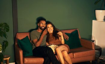 A couple sits on a couch while watching a movie and eating popcorn.