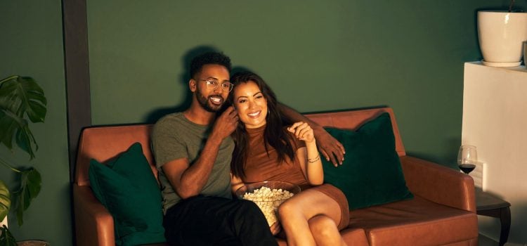 A couple sits on a couch while watching a movie and eating popcorn.