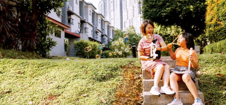 A mom and a boy sit outside in the garden. The mom holds a black and white cat while the boy blows bubbles.