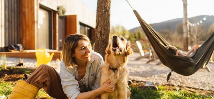 A woman sits outside a house and pets her golden retriever. Nearby a person lounges on a hammock.