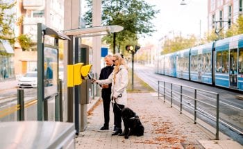 A blind woman uses a keypad outside of a train station while a man stands by her side.