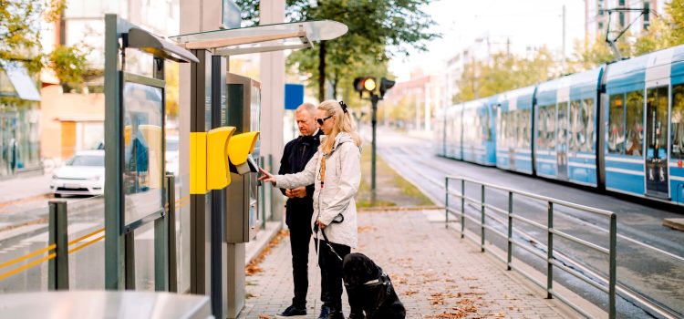 A blind woman uses a keypad outside of a train station while a man stands by her side.
