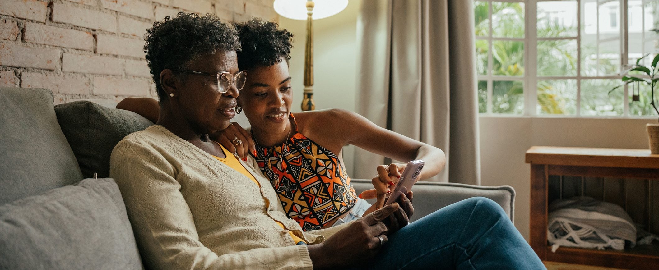 A young woman points something out on her mother’s phone as they sit together on the couch. 
