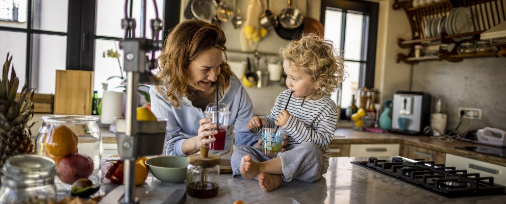 A woman stands in her kitchen holding a drink with straw, next to her young daughter who sits on the counter.