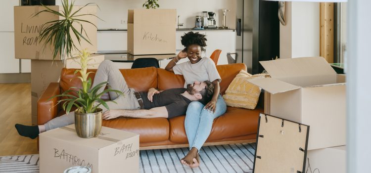 A couple relaxes on a couch surrounded by unpacked moving boxes in their living room.
