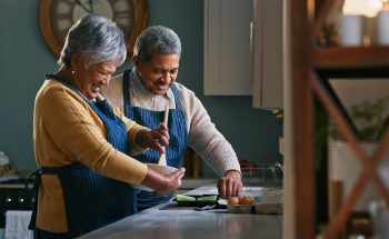 A retired couple smiles while cooking together in their kitchen.