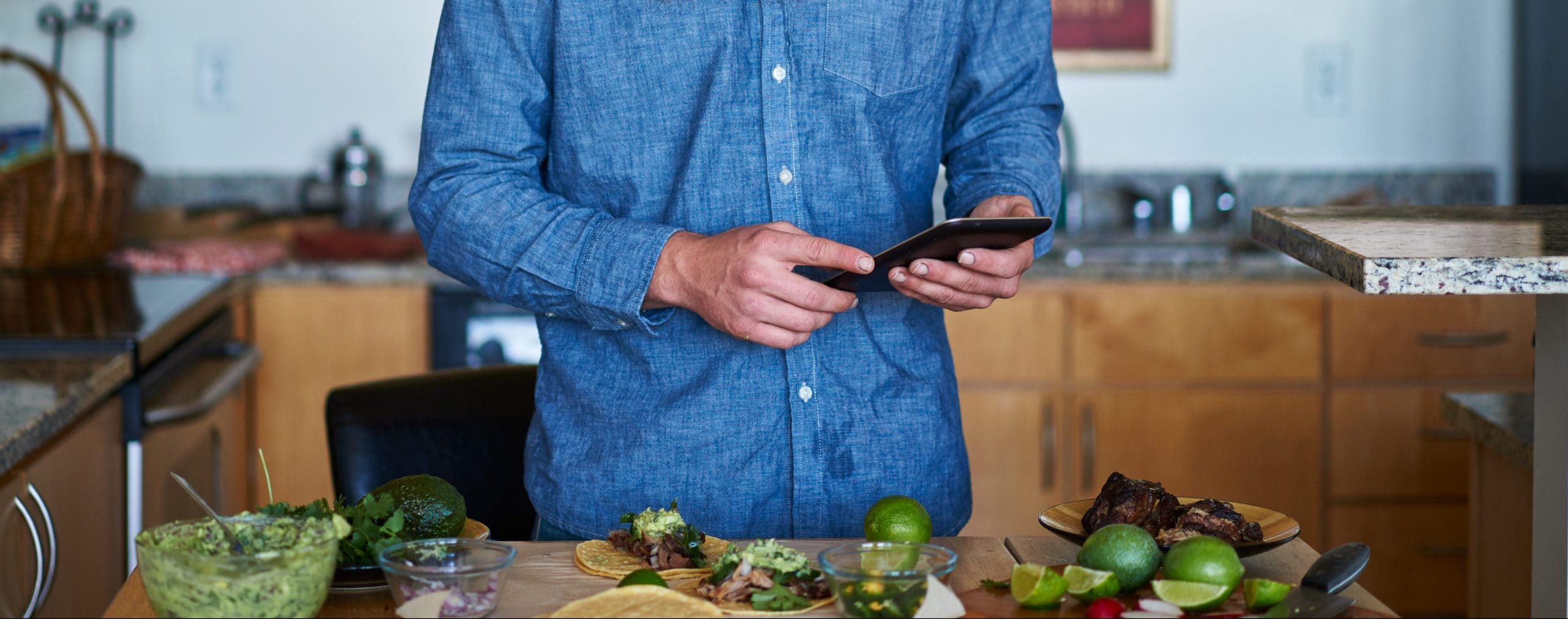 A man standing in his kitchen, making tacos and looking at something on his phone.