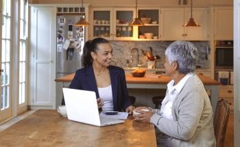 A woman discusses finances with an advisor at her kitchen table, with documents and a laptop in front of them.