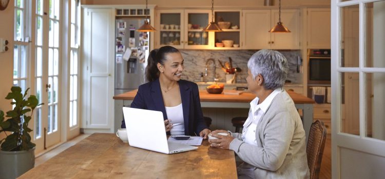A woman discusses finances with an advisor at her kitchen table, with documents and a laptop in front of them.