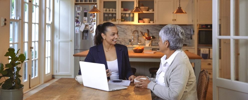 A woman discusses finances with an advisor at her kitchen table, with documents and a laptop in front of them.