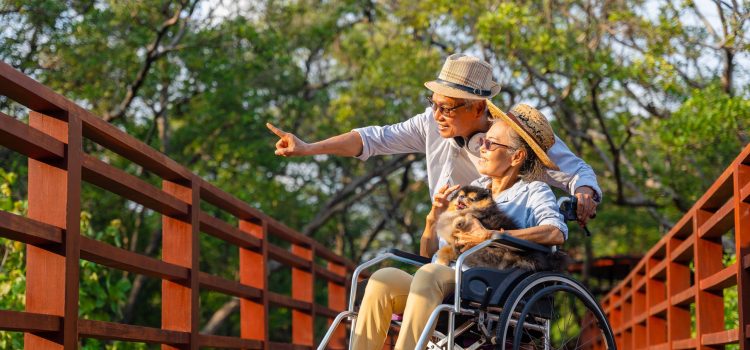 Older man points to something outside while on a walk with his wife who is in a wheelchair holding a dog.