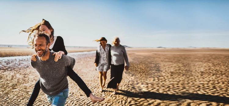 A man carries a woman on piggyback while walking on the beach, while two women walk behind with their arms around each other.
