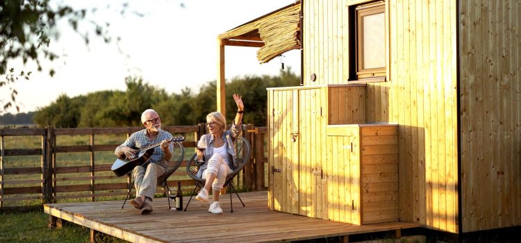 A retired couple sits on chairs outside. The man plays a guitar while the woman sings and drinks wine.
